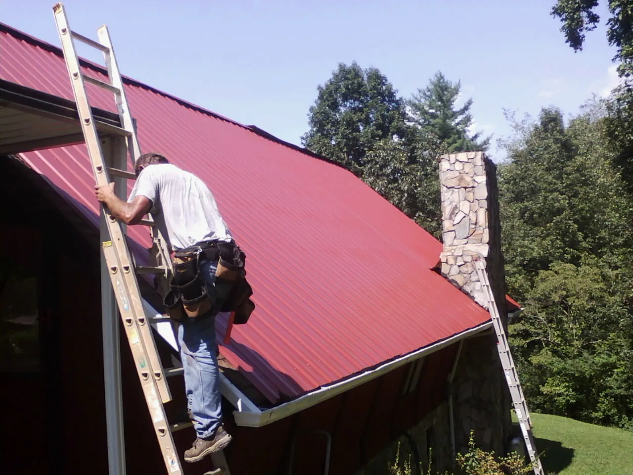 Roofer on a ladder beside a red metal roof and stone chimney — Soffit & Fascia Repair by Roofing Brothers in Buffalo